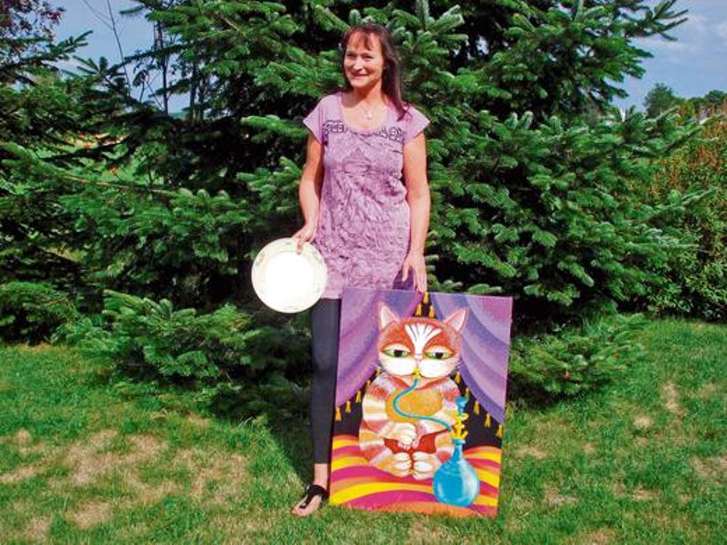 Handwerklich perfekt, inhaltlich heiter bis lieblich: Ruth Wunderlich mit ihrem neusten Gemälde, das eine bekiffte Katze zeigt, und einem Teller, den sie als Designerin dekorierte. Foto: asz
